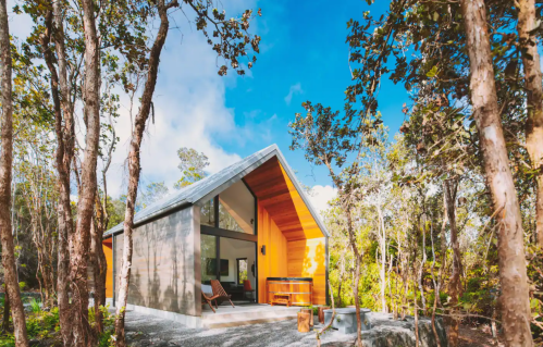 A modern cabin with a sloped roof nestled among trees, featuring large windows and a cozy outdoor seating area.