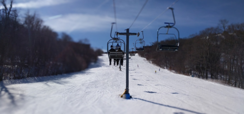 Ski lift carrying passengers over a snowy slope under a clear blue sky. Trees line the background.