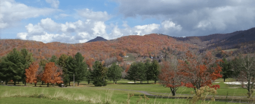 A scenic view of colorful autumn trees on rolling hills under a partly cloudy sky.