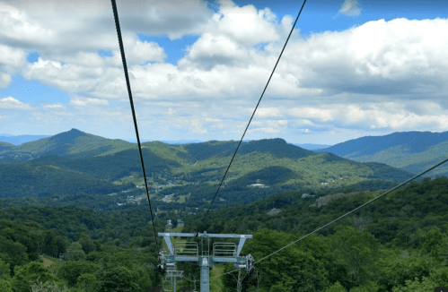 A scenic view from a chairlift, overlooking lush green mountains and a blue sky with fluffy clouds.