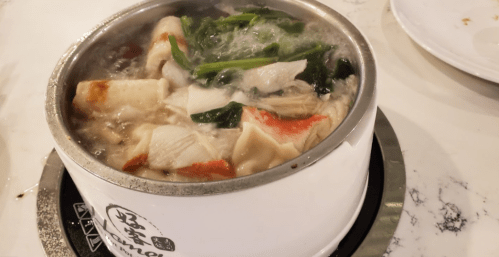 A steaming pot of soup with fish, vegetables, and herbs, served in a white bowl on a heating element.
