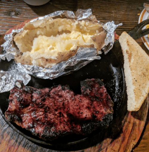 A plate with a grilled steak, a baked potato with butter, and a slice of bread on a wooden surface.