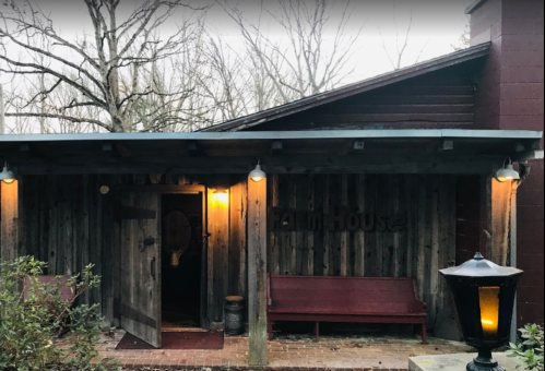 A rustic wooden building with a sign reading "Tavern House," featuring a bench and lanterns, surrounded by trees.