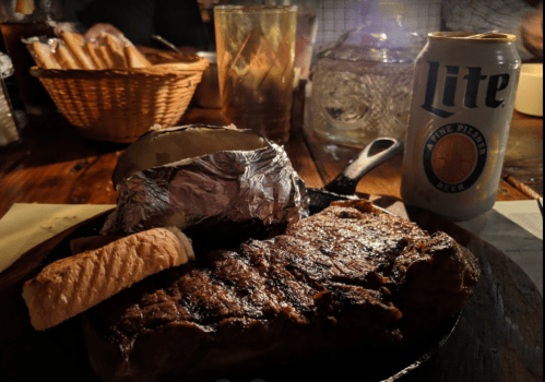 A sizzling steak on a cast iron skillet, with a baked potato, bread roll, and a can of beer in a dimly lit setting.