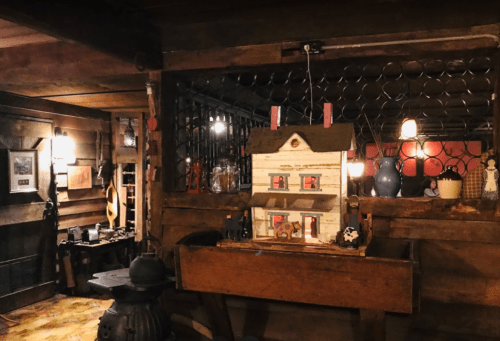 A cozy, rustic interior featuring a wooden dollhouse, warm lighting, and various vintage decor elements.