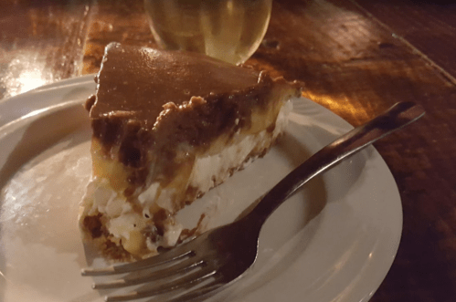 A slice of layered dessert on a plate, featuring chocolate, cream, and banana, with a fork beside it.