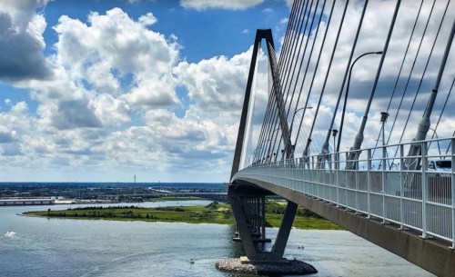 A modern cable-stayed bridge spans a river under a blue sky with fluffy clouds, surrounded by greenery and water.