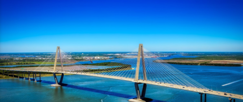 Aerial view of a cable-stayed bridge spanning a blue river under a clear sky, with green land in the background.