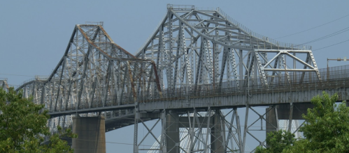 A close-up view of a large, weathered metal bridge with a blue sky in the background and greenery in the foreground.