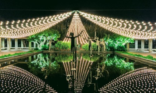 A serene night scene with a pond reflecting a canopy of lights and sculptures surrounded by lush greenery.