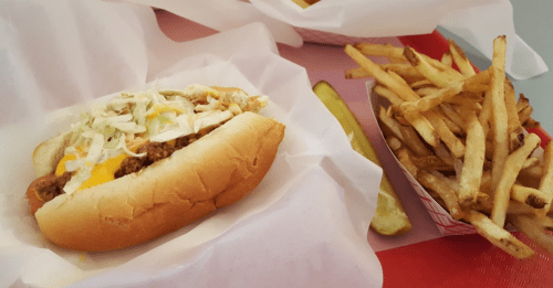 A hot dog topped with lettuce and cheese, served with a side of crispy French fries and a pickle slice.
