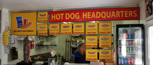 A colorful hot dog stand interior with a menu featuring various hot dog options and a staff member behind the counter.