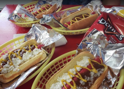 Four hot dogs in yellow baskets, topped with mustard and chili, alongside a bag of Doritos.