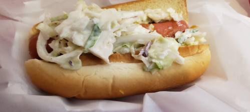 A hot dog in a bun topped with creamy coleslaw, served on white paper.
