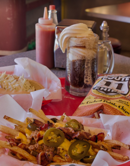A plate of loaded fries topped with cheese, bacon, and jalapeños, alongside a root beer float and chips.