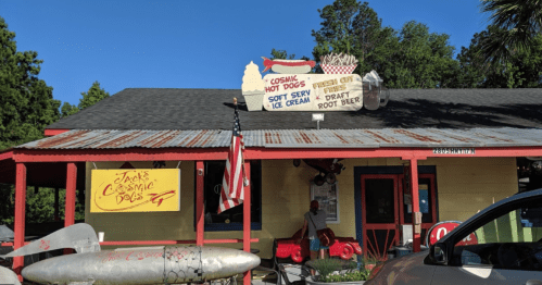 A colorful roadside eatery with a sign for hot dogs and ice cream, featuring a vintage car and an American flag.