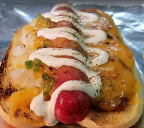 A toasted hot dog in a bun topped with melted cheese, diced onions, jalapeños, and drizzled with sour cream.