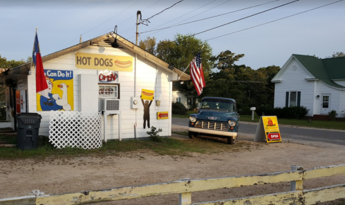 A small hot dog stand with a vintage truck parked outside, featuring flags and colorful signage.
