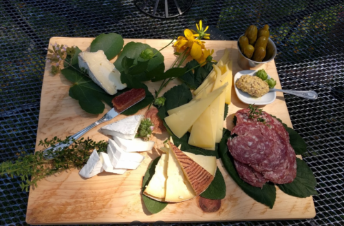 A wooden platter with various cheeses, cured meats, pickles, mustard, and fresh flowers on a textured surface.