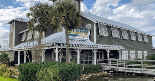 A wooden building with a sign reading "Alligator Adventure," surrounded by greenery and water under a cloudy sky.