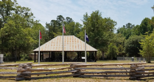 A rustic pavilion with a rusty roof, surrounded by trees and a wooden fence, featuring flags on poles.