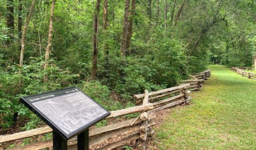 A wooded path with a split-rail fence and an informational sign about the area, surrounded by lush greenery.