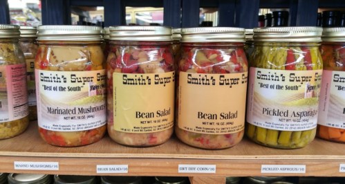 Jars of various pickled vegetables on a shelf, including marinated mushrooms, bean salad, and pickled asparagus.