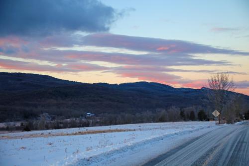 A snowy landscape at dusk, with mountains in the background and colorful clouds in the sky. A road leads into the scene.