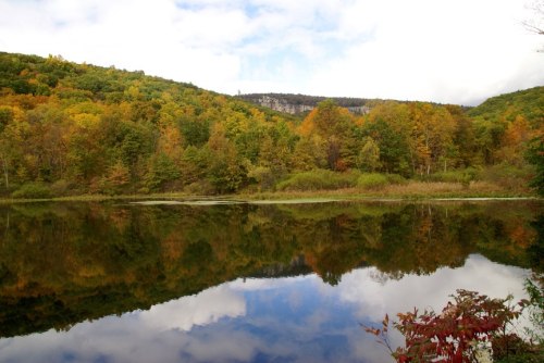 A serene lake reflects colorful autumn trees and a cloudy sky, surrounded by lush hills.