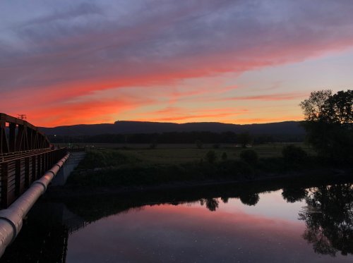 A vibrant sunset reflects on a calm river, with silhouettes of hills and a bridge in the foreground.