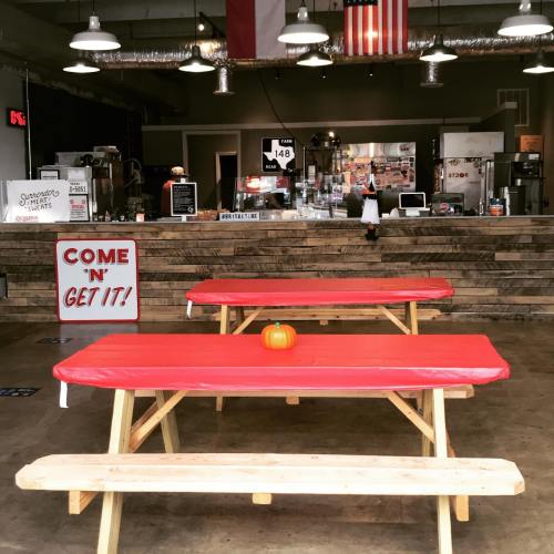 A cozy eatery with wooden tables, red tablecloths, and a pumpkin centerpiece, featuring a counter and flags in the background.