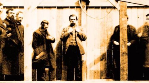 A historical black-and-white photo of a group of men, some standing, others partially obscured, in front of a wooden backdrop.