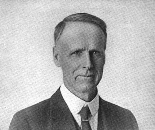 Black and white portrait of a man in a suit with short hair, looking directly at the camera.