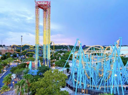 A vibrant amusement park scene featuring colorful roller coasters and a tall drop tower under a cloudy sky.