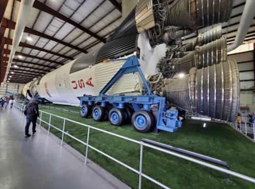 A large rocket engine displayed indoors, with a blue transport vehicle beside it and visitors in the background.
