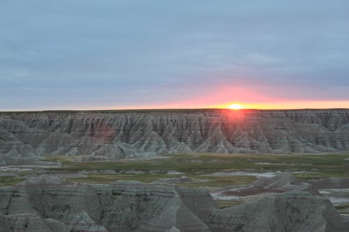 Sunrise over rugged, layered rock formations in a vast, open landscape under a cloudy sky.