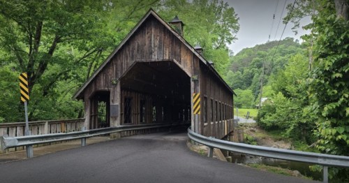 A rustic wooden covered bridge surrounded by lush green trees and a winding road.