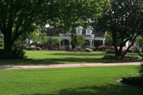 A large, elegant house surrounded by lush greenery and colorful flowers, set on a spacious lawn.
