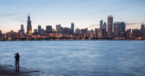 A silhouette of a person photographing the Chicago skyline at dusk, with water in the foreground and city lights glowing.