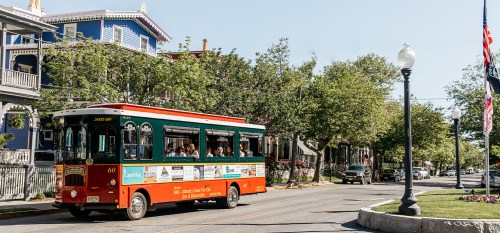 A colorful trolley bus drives down a tree-lined street with historic buildings and parked cars in the background.