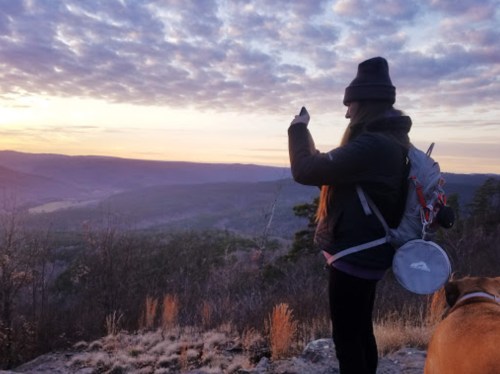 A person in a beanie takes a photo of a sunset while standing on a rocky ledge with a dog nearby.