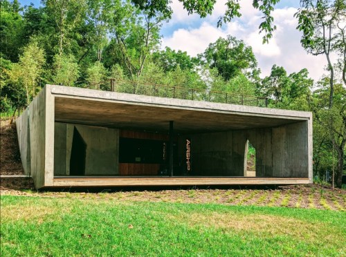 A modern concrete building with large openings, set in a green landscape surrounded by trees.
