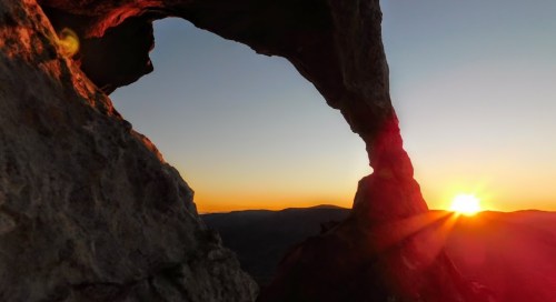 Sunset view through a rocky arch, with warm colors illuminating the landscape and mountains in the background.