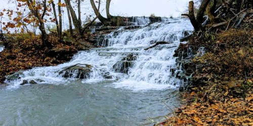 A serene waterfall cascades over rocky steps, surrounded by autumn foliage and a calm pool of water below.