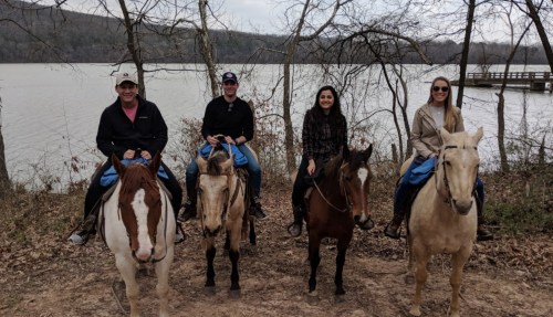 Four people on horseback by a lake, surrounded by trees, smiling at the camera.