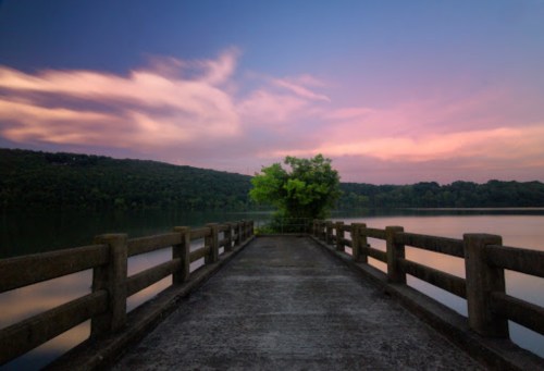 A serene pier extends into a calm lake, surrounded by lush greenery and a colorful sunset sky.