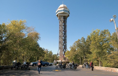 A tall observation tower surrounded by trees, with people and motorcycles parked nearby on a clear day.