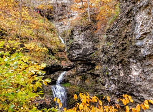 A scenic waterfall cascades through rocky terrain, surrounded by vibrant autumn foliage in a forested area.