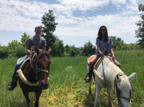 Two people riding horses in a grassy field under a partly cloudy sky.