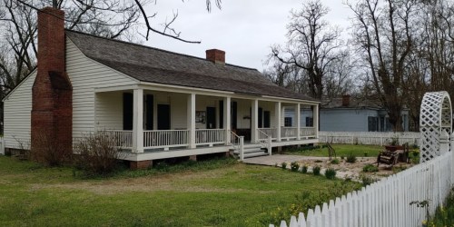A historic white house with a porch, surrounded by trees and a garden, set against a cloudy sky.